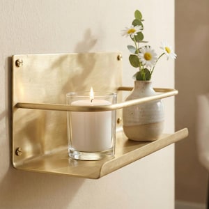 May include: A brass wall shelf with a lit candle in a glass and a small vase with daisies. The shelf is mounted on a beige wall. The candle flame is bright, and the vase is a neutral color.
