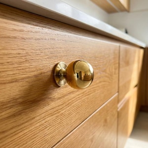 Peut inclure: Gros plan sur une armoire en bois avec un bouton sphérique doré. L'armoire présente une finition en grain de bois brun clair. Le bouton est rond avec une base circulaire. L'arrière-plan montre un comptoir blanc et d'autres armoires en bois.