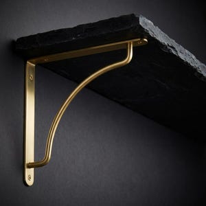 May include: A black slate shelf supported by a gold-colored metal bracket. The bracket has a curved design and is attached to the shelf and wall with screws. The shelf has a rough, natural edge.