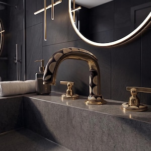 May include: A gold-toned bathroom faucet with a curved spout and two matching handles, featuring a decorative black and white circular pattern inlay on the base and spout. The faucet is mounted on a gray stone countertop next to a soap dispenser.