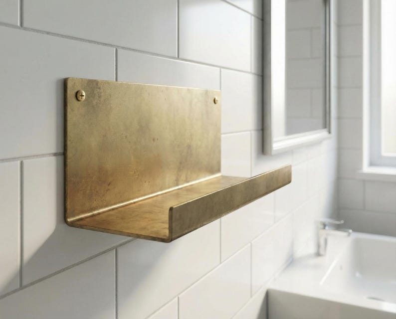 May include: A brass-colored wall-mounted shelf with a rectangular back and a horizontal shelf. The shelf is attached to a white tiled wall with two screws. The shelf is in a bathroom setting.