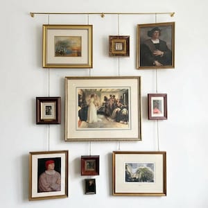 Könnte beinhalten: Eine Sammlung gerahmter Kunstwerke, darunter Gemälde und Drucke, die an einer Messingstange mit Ketten aufgehängt sind. Die Kunstwerke zeigen verschiedene Motive, darunter Landschaften und Porträts, mit verzierten goldenen und braunen Rahmen.