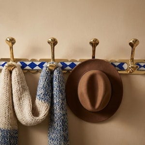 May include: A brass wall-mounted coat rack with four hooks, a blue and white patterned accent, and a brown fedora hat. Two knitted scarves, one beige and blue, the other blue, hang from the hooks.