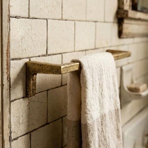 May include: A brass towel bar mounted on a tiled wall. A folded white and beige striped towel hangs from the bar. The wall tiles are off-white with a cracked, aged appearance, suggesting a vintage aesthetic.