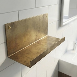 May include: A wall-mounted shelf made of brass. The shelf has a rectangular back and a horizontal ledge. Two screws secure the shelf to the white tiled wall. The shelf is in a bathroom setting, with a mirror and sink visible in the background.