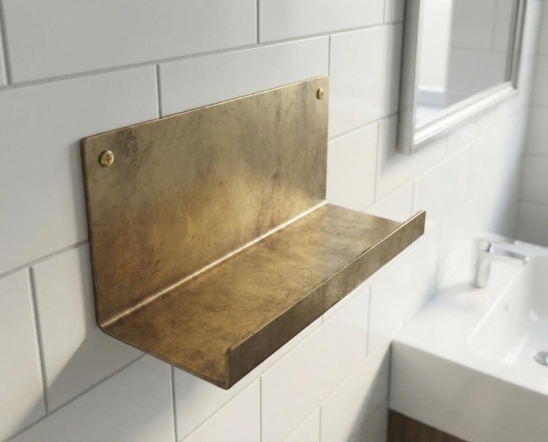 May include: A wall-mounted shelf made of brass. The shelf has a rectangular back and a horizontal ledge. Two screws secure the shelf to the white tiled wall. The shelf is in a bathroom setting, with a mirror and sink visible in the background.