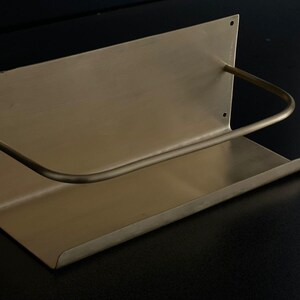 May include: A brass-toned wall-mounted shelf with a curved bar. The shelf has a rectangular base and a back panel with three small holes. The curved bar extends from the back panel to the front of the shelf.