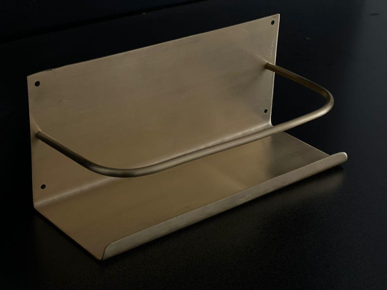May include: A brass-toned wall-mounted shelf with a curved bar. The shelf has a rectangular base and a back panel with three small holes. The curved bar extends from the back panel to the front of the shelf.