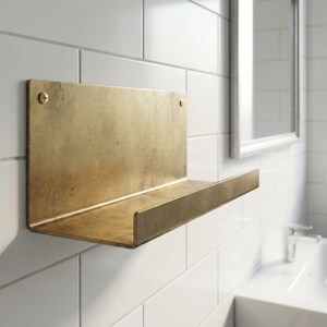 May include: A brass-colored metal shelf mounted on a white tiled wall. The shelf has a rectangular back and a horizontal ledge. Two brass screws secure the shelf to the wall. The bathroom fixture is near a sink and mirror.