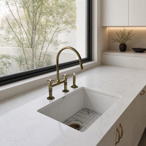 Puede incluir: Un área de fregadero de cocina con una cubeta rectangular blanca, una rejilla metálica en el fondo y un grifo de latón con dos mandos en cruz y un rociador lateral, instalado sobre una encimera de mármol blanco con vetas grises.