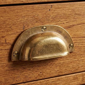 May include: Close-up of a brass drawer pull with a semi-circular design, mounted on a wooden drawer. The pull is secured with three screws and has a weathered, antique appearance. The wood grain is visible.