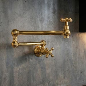 May include: A gold-colored, wall-mounted pot filler faucet with a cross-shaped handle and a ball joint design. The faucet has a polished finish and is mounted on a textured gray wall.