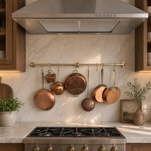 May include: A kitchen scene featuring copper cookware hanging from a brass pot rack above a gas stove. The copper pots and pans vary in size and shape, reflecting a classic kitchen aesthetic. A stainless steel range hood is mounted above the stove.