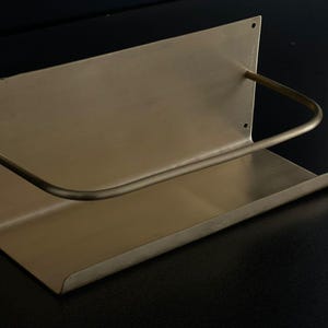 May include: A brass-toned wall-mounted shelf with a curved bar. The shelf has a rectangular base and a back panel with three small holes. The curved bar extends from the back panel to the front of the shelf.