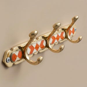 Puede incluir: Un perchero de pared dorado con tres ganchos curvos. El perchero presenta incrustaciones decorativas de esmalte en forma de rombos naranjas y blancos. Los ganchos están montados en una barra rectangular con detalles decorativos.