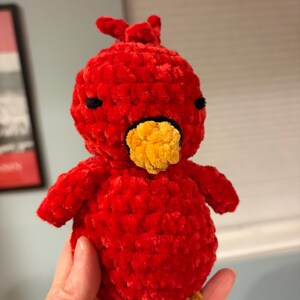 May include: A handmade, plush red bird with a yellow beak and feet. The bird has small black eyes and a tuft of feathers on its head. The soft, textured material gives it a cuddly appearance, perfect for a child's toy or a decorative item.