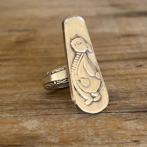 May include: A silver-toned ring with a rectangular face featuring an embossed design of a bird-like creature. The ring has a detailed band with an ornate pattern. The ring is set against a wooden surface.