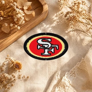 Pode incluir: Um emblema bordado de formato oval com o logótipo dos San Francisco 49ers. O design consiste num monograma SF branco entrelaçado centrado num fundo oval vermelho, rodeado por uma margem dourada fina e um rebordo exterior preto espesso.
