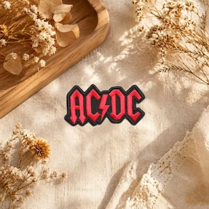 May include: An embroidered patch featuring the AC/DC logo in red lettering with a black border. The letters are stylized with sharp, angular edges, and a lightning bolt symbol separates the two parts of the band name in the center of the design.