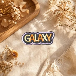 Può includere: Una toppa ricamata con sfondo nero e bordo viola chiaro. La parola GALAXY è cucita al centro con filo che alterna giallo e arancione, caratterizzata da una forma ondulata simile a una nuvola che segue il contorno del testo.