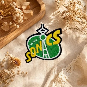 Puede incluir: Un parche bordado con forma ovalada verde y borde blanco. La palabra SONICS está cosida en letras amarillas sobre el fondo verde, con un gráfico blanco de la torre Space Needle integrado en el diseño del parche.