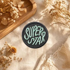 以下が含まれることがあります： 黒い背景に明るいミントグリーンの糸でSUPERSTARという文字が刺繍された円形のワッペン。ワッペンには暗い色の縁取りがあり、文字がはっきりと読み取れる平らな円形のテキスタイルアイテムとして表示されています。