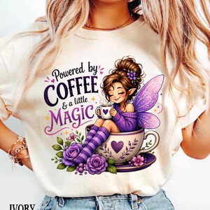 May include: An ivory t-shirt featuring a graphic of a fairy with brown hair and purple wings sitting in a large teacup. The text reads Powered by COFFEE & a little MAGIC. The design includes purple roses, striped socks, and a small cup with a heart.