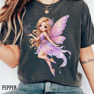 May include: A charcoal gray t-shirt featuring a graphic illustration of a blonde fairy with large, translucent purple wings. The fairy wears a purple and pink petal-style dress and is surrounded by small sparkling dust particles. The word PEPPER is printed.