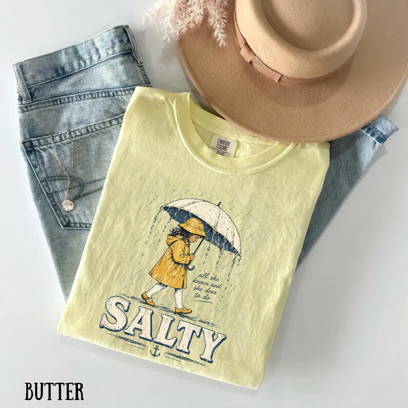 May include: A pale yellow t-shirt featuring a graphic of a girl in a yellow raincoat and hat under an umbrella in the rain. The word "SALTY" is printed below the graphic. Blue jeans and a beige hat are also visible.