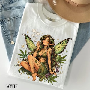 May include: A white t-shirt featuring a graphic illustration of a fairy with long brown hair and green wings. The fairy is wearing a green top and skirt, holding a lit cigarette, and is surrounded by cannabis leaves, small white flowers, and stars.