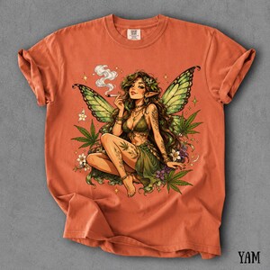 May include: A burnt orange short-sleeve t-shirt featuring a graphic print of a fairy with green wings, long brown hair, and a green outfit. The fairy is surrounded by cannabis leaves, small white flowers, and smoke, holding a lit cigarette.