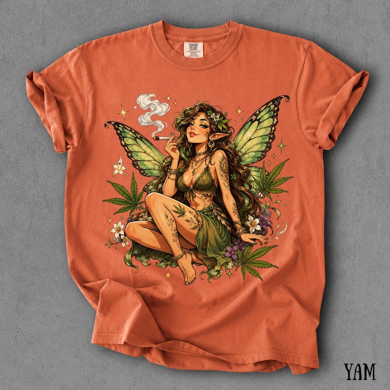 May include: A burnt orange short-sleeve t-shirt featuring a graphic print of a fairy with green wings, long brown hair, and a green outfit. The fairy is surrounded by cannabis leaves, small white flowers, and smoke, holding a lit cigarette.