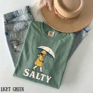 May include: A light green t-shirt featuring a graphic of a girl in a yellow raincoat holding an umbrella, with the word "SALTY" below. The shirt is styled with acid wash jeans and a tan hat. The shirt is a size L/G.