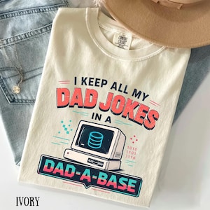 Può includere: T-shirt color avorio con la scritta "I KEEP ALL MY DAD JOKES IN A DAD-A-BASE" in rosso e turchese. La grafica include un computer retrò con un'icona di database. La maglietta è in materiale Comfort Colors.