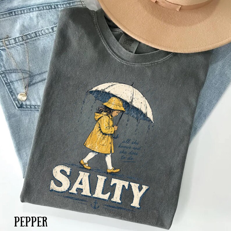 May include: A grey t-shirt featuring a graphic of a girl in a yellow raincoat and hat under an umbrella. The word "SALTY" is printed below the image. A tan hat and blue jeans are in the background. The text "all she knows and she does to do" is also visible.