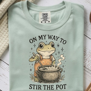 Puede incluir: Camiseta verde mar con una rana de dibujos animados removiendo una olla, el texto dice "ON MY WAY TO STIR THE POT". La camiseta tiene una etiqueta Comfort Colors y una pegatina "Fast Delivery".