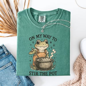 Puede incluir: Una camiseta verde claro con el texto "ON MY WAY TO STIR THE POT" y una rana de dibujos animados removiendo una olla. La camiseta está doblada y tiene un collar plateado encima. Unos vaqueros azules y unos zapatos blancos también están en la imagen.