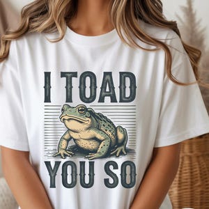 May include: White t-shirt with a green and yellow toad graphic and the text "I TOAD YOU SO". The design features a distressed font and horizontal lines. The tee has a crew neck and short sleeves.