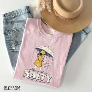May include: A pale pink t-shirt featuring a graphic of a girl in a yellow raincoat and hat under an umbrella in the rain. The word "SALTY" is printed below. A tan hat and blue jeans are also visible.