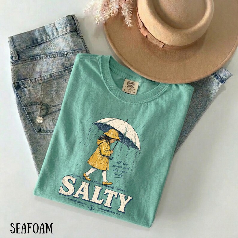 May include: Seafoam green t-shirt featuring a graphic of a girl in a yellow raincoat holding an umbrella, with the word "SALTY" below. The shirt is styled with acid wash jeans and a tan hat.