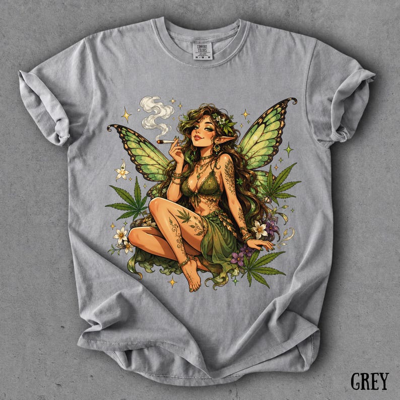May include: A light grey t-shirt featuring a graphic illustration of a fairy with green wings, long brown hair, and pointed ears. The fairy is sitting among cannabis leaves and small white flowers, holding a lit cigarette with smoke rising.