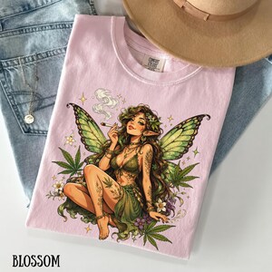 May include: A light pink t-shirt featuring a graphic illustration of a fairy with green wings, long brown hair, and tattoos, surrounded by cannabis leaves and flowers. The fairy is holding a smoking cigarette. The shirt has a crew neckline.