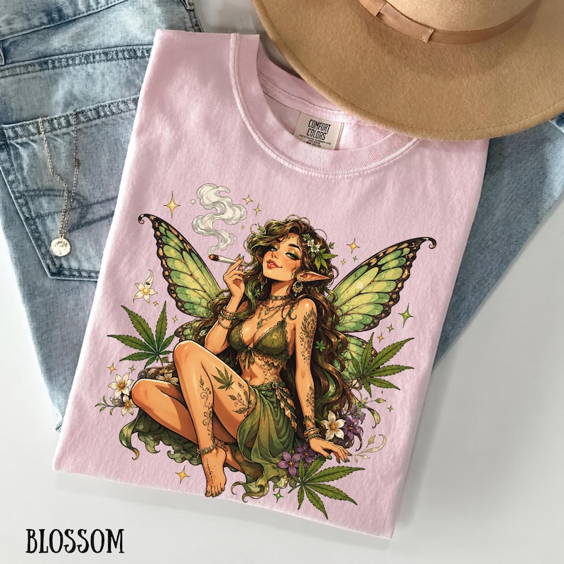 May include: A light pink t-shirt featuring a graphic illustration of a fairy with green wings, long brown hair, and tattoos, surrounded by cannabis leaves and flowers. The fairy is holding a smoking cigarette. The shirt has a crew neckline.