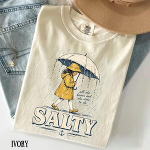 May include: Ivory t-shirt featuring a graphic of a child in a yellow raincoat and hat under an umbrella in the rain. The word "SALTY" is printed in blue, with an anchor below. Text on the shirt reads "all she knows and she does to do."