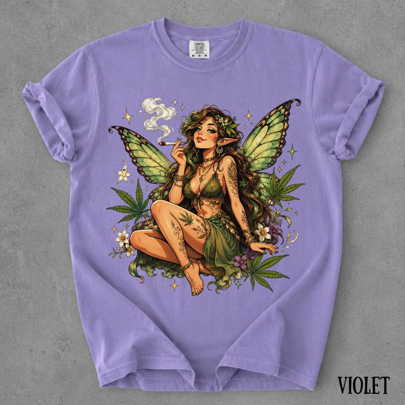 May include: A light purple t-shirt featuring a graphic illustration of a fairy with green wings, long brown hair, and a green outfit. The fairy is holding a lit cigarette, surrounded by cannabis leaves, small white flowers, and yellow stars.