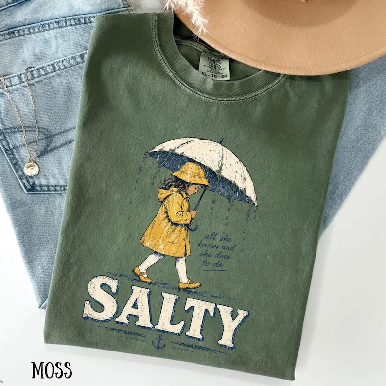 May include: A moss green t-shirt featuring a graphic of a girl in a yellow raincoat and hat under an umbrella in the rain. The word "SALTY" is printed in large letters below the illustration. The shirt is on top of blue denim jeans.