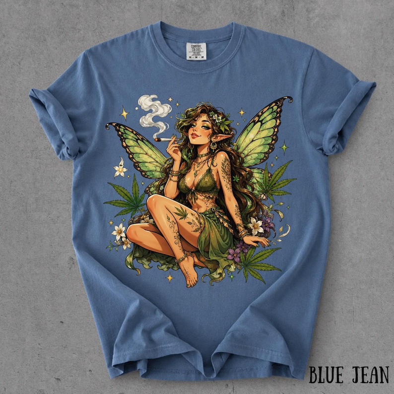 May include: A blue crew neck t-shirt featuring a graphic illustration of a fairy with green wings, long brown hair, and a green outfit. The fairy is holding a lit cigarette with smoke rising, surrounded by green cannabis leaves and white flowers.
