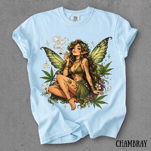 May include: A light blue t-shirt featuring a graphic illustration of a fairy with green wings, long brown hair, and a green outfit. The fairy is holding a lit cigarette, surrounded by cannabis leaves, small white flowers, and sparkling stars.