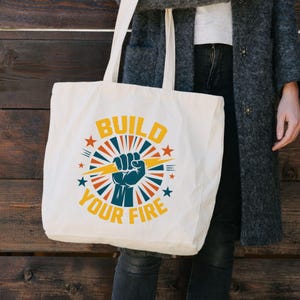 Könnte beinhalten: Cremefarbene Tragetasche mit dem Text "BUILD YOUR FIRE" in Gelb und Marineblau. Das Design umfasst eine geballte Faust, einen Blitz und Sternenstrahlen-Elemente in Rot, Blau und Gelb. Die Tasche hat lange Henkel.