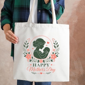Puede incluir: Bolso tote de color crema con una silueta verde de madre e hijo dentro de una corona floral. Las palabras "Happy Mother's Day" están impresas debajo en una fuente cursiva, con pequeños corazones y elementos decorativos.